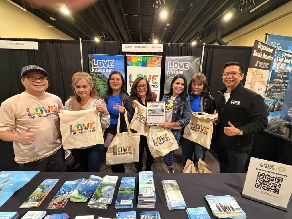 Group of people posing at a travel expo booth, holding tote bags that say 'LOVE THE PHILIPPINES' with colorful banners in the background.