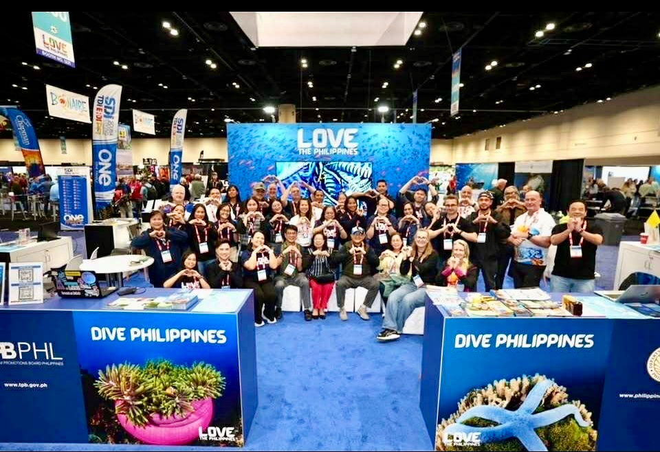 Large group photo at a trade show booth for Dive Philippines, with a blue backdrop reading 'Love the Philippines' and coral imagery on the counters.